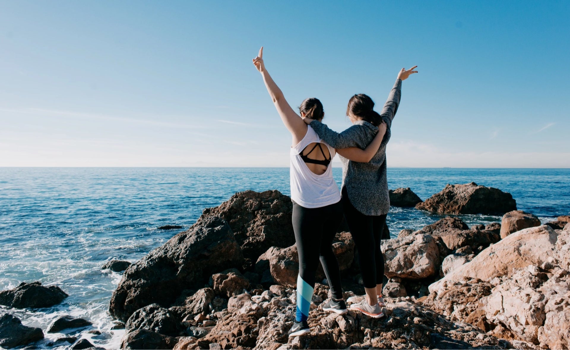 Two people standing on rocks by the ocean with arms raised, enjoying the view.